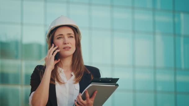 一位身穿正装的年轻女工程师或建筑师 带着建筑蓝图站在蓝色窗户的大楼旁边 在电话上交谈 — 图库视频影像