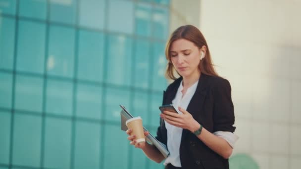 成功的女商人穿着西装 走在一个现代玻璃商务区附近 拿着咖啡杯 使用智能手机 新业务 工作日 自由职业者 — 图库视频影像