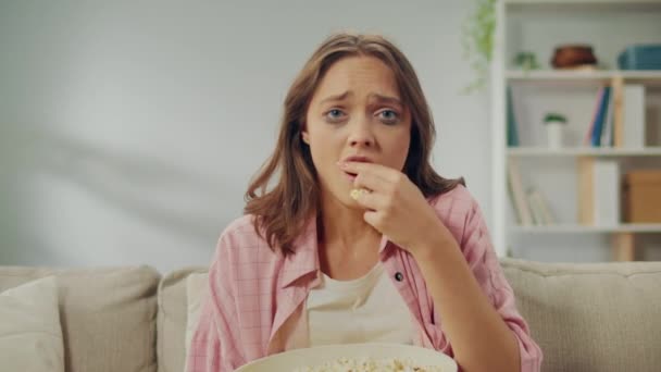 一个年轻的女人坐在沙发上 吃爆米花 看悲伤的电影 女人一边看情景剧一边哭 电影作为一种情感发泄和自我表达的方式 电影及其角色 — 图库视频影像