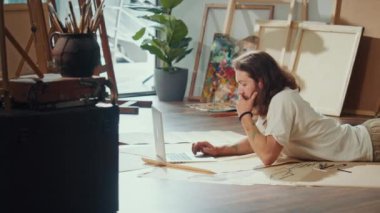 Bir Sanatçı Parlak Stüdyosunda Yerde Otururken Yeni Sanatlar Oluşturmak İçin Resim Kullanıyor. Eserleri ile çevrelenmiş olarak Endişeleri yönetmek ve zihinsel sağlığı geliştirmek için Sakin Kullanım Sanatı Odaklanıyor.