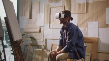 Bir Erkek Sanatçı Parlak Stüdyosunda Bitiş Çalışmaları ile çevrelenmiş eşsiz bir sanat yaratmak için VR gözlükleri kullanır. Video, 3D Sanat Yaratıcılığı için Geleneksel Boyama ve VR Teknolojisinin Harmanını Vurguluyor.