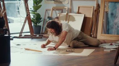 Genç Bir Sanatçı Samimi Stüdyosunda Yerde Boya Yapıyor, Etrafı Bitirdiği Çalışmalarla çevrili. Parlak, Yaratıcı Uzay, ona yeni, eşsiz sanat eserlerini sıcak ve davetkar bir ortamda yaratması için ilham veriyor..
