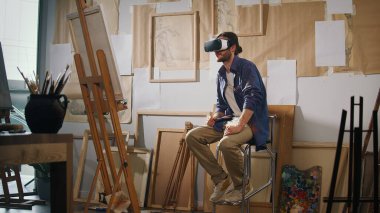 Bir Erkek Sanatçı Parlak Stüdyosunda Bitiş Çalışmaları ile çevrelenmiş eşsiz bir sanat yaratmak için VR gözlükleri kullanır. Video, 3D Sanat Yaratıcılığı için Geleneksel Boyama ve VR Teknolojisinin Harmanını Vurguluyor.