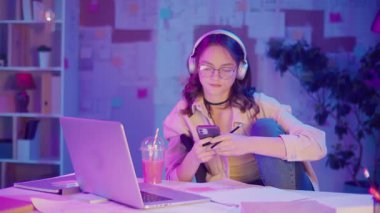 Gen Z Yaşam Tarzı, Dijital Çoklu Görev, Neon Mood. Genç Kız Müzik Dinlerken ve Dizüstü bilgisayar izlerken resim çizer. Neon Işığında Modern Gençliğin Yaratıcı Kaosu 'nda Kişisel İfade