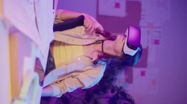 Dikey Video Karışık Gerçeklik, Immersive Teknoloji, Gen Z Geleceği. Neon-Lit Room 'da oturan kız VR ya da AR gözlükleri takıyor, Dijital Alanla etkileşimde. Görsel olarak Yoğun Sanal Dünya Kişisel Anı