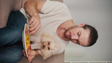 Dikey video. Basit Mutluluklar, Samimi Anlar, Günlük Bond. Genç adam köpeği ile koltukta oturur, gevşemiş zamanı paylaşır ve patates kızartması yer. Dürüst, Sıcak Dostluk Sahnesi, Yemek ve Evde Rahatlık