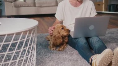 Yumuşak İş Arkadaşı, Duygusal Odaklanma, Uzaktan Çalışma. Adam kanepeye oturur ve dizüstü bilgisayarı üzerinde çalışır ve köpeğiyle etkileşim içindedir. Sakin, neşeli bir Denge, Bağlanma ve Evcil Hayvanlarla Modern Yaşam