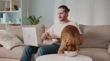 Sağlıklı rutin, Evcil Hayvan Besleme, Dijital Denge. Köpeği yakınlarda kendi yemeğinin tadını çıkarırken adam masa başında çalışıyor ve yiyor. Sakin, Senkronize Sağlık ve Günlük Odaklanma