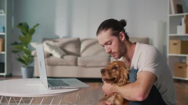 Yumuşak İş Arkadaşı, Duygusal Odaklanma, Uzaktan Çalışma. Adam kanepeye oturur ve dizüstü bilgisayarı üzerinde çalışır ve köpeğiyle etkileşim içindedir. Sakin, neşeli bir Denge, Bağlanma ve Evcil Hayvanlarla Modern Yaşam