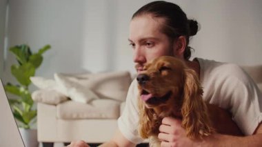 Mikro neşeler, Akıl Sağlığı, Günlük Denge. Genç Adam, köpeği ile koltukta iletişim kurarken dizüstü bilgisayarda çalışıyor. Yumuşak, destekleyici bir an Harmanlayıcı Odak, Duygu ve Bağlantı