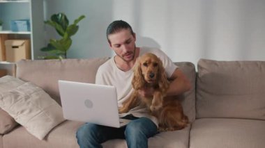 Yumuşak İş Arkadaşı, Duygusal Odaklanma, Uzaktan Çalışma. Adam kanepeye oturur ve dizüstü bilgisayarı üzerinde çalışır ve köpeğiyle etkileşim içindedir. Sakin, neşeli bir Denge, Bağlanma ve Evcil Hayvanlarla Modern Yaşam