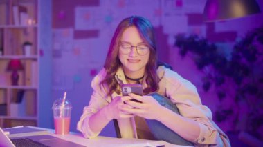 Dijital alışkanlıklar, Gen Z Yaşam Tarzı, Duygusal Odaklanma. Genç Kadın Neon Odasında Oturuyor, Telefonuna Sosyal Medya Yazıyor. Yalnızlık, Yansıma veya Ekran Kültüründe Kaçış Estetik Anı