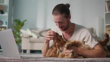 Mikro neşeler, Akıl Sağlığı, Günlük Denge. Genç Adam, köpeği ile koltukta iletişim kurarken dizüstü bilgisayarda çalışıyor. Yumuşak, destekleyici bir an Harmanlayıcı Odak, Duygu ve Bağlantı