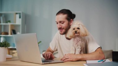 Mikro neşeler, Akıl Sağlığı, Günlük Denge. Genç Adam, köpeği ile koltukta iletişim kurarken dizüstü bilgisayarda çalışıyor. Yumuşak, destekleyici bir an Harmanlayıcı Odak, Duygu ve Bağlantı