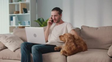 Yumuşak İş Arkadaşı, Duygusal Odaklanma, Uzaktan Çalışma. Adam kanepeye oturur ve dizüstü bilgisayarı üzerinde çalışır ve köpeğiyle etkileşim içindedir. Sakin, neşeli bir Denge, Bağlanma ve Evcil Hayvanlarla Modern Yaşam