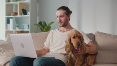 Yumuşak İş Arkadaşı, Duygusal Odaklanma, Uzaktan Çalışma. Adam kanepeye oturur ve dizüstü bilgisayarı üzerinde çalışır ve köpeğiyle etkileşim içindedir. Sakin, neşeli bir Denge, Bağlanma ve Evcil Hayvanlarla Modern Yaşam
