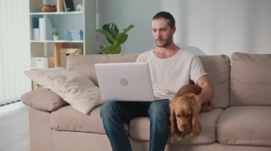 Yumuşak İş Arkadaşı, Duygusal Odaklanma, Uzaktan Çalışma. Adam kanepeye oturur ve dizüstü bilgisayarı üzerinde çalışır ve köpeğiyle etkileşim içindedir. Sakin, neşeli bir Denge, Bağlanma ve Evcil Hayvanlarla Modern Yaşam