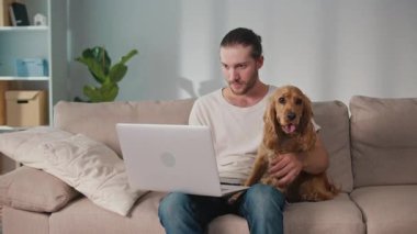 Yumuşak İş Arkadaşı, Duygusal Odaklanma, Uzaktan Çalışma. Adam kanepeye oturur ve dizüstü bilgisayarı üzerinde çalışır ve köpeğiyle etkileşim içindedir. Sakin, neşeli bir Denge, Bağlanma ve Evcil Hayvanlarla Modern Yaşam