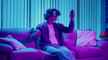 Young Man Sits on Sofa at Night in AR Glasses, Interacting with Virtual Interface. Immersive Experience in Digital World of Tomorrow. Mixed Reality, Future Technology, Mental Escape.