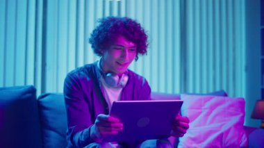 Genç adam geceleri koltukta oturur, Tablet ekranına güler. Dijital Günlük Yaşamda Eğlence, Rahatlama ve Bağlanma Anı. Ruh Sağlığı, Dijital Bağlantı, Duygusal Rahatlama.