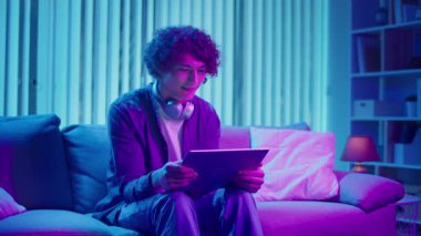 Genç adam geceleri koltukta oturur, Tablet ekranına güler. Dijital Günlük Yaşamda Eğlence, Rahatlama ve Bağlanma Anı. Ruh Sağlığı, Dijital Bağlantı, Duygusal Rahatlama.