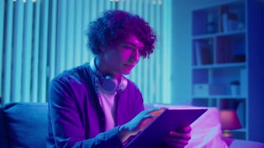 Genç adam tabletle koltukta oturuyor, konsantrasyonla çalışıyor. Esnek Teknolojik Çalışma Akışı 'nda sakin ve etkili bir akşam. Odaklanma Kipi, Dijital Üretim, Özyönetim.
