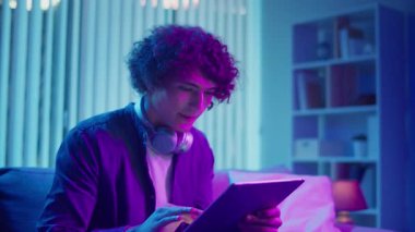 Genç adam tabletle koltukta oturuyor, konsantrasyonla çalışıyor. Esnek Teknolojik Çalışma Akışı 'nda sakin ve etkili bir akşam. Odaklanma Kipi, Dijital Üretim, Özyönetim.