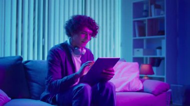 Genç adam tabletle koltukta oturuyor, konsantrasyonla çalışıyor. Esnek Teknolojik Çalışma Akışı 'nda sakin ve etkili bir akşam. Odaklanma Kipi, Dijital Üretim, Özyönetim.