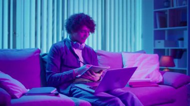 Genç Adam Neon Light 'ta, Kanepede Gece Çalışır, Not Defteri ve Laptop Tutar. Sakin Üretim ve Ruh hali Işığı ile Gen Z Çalışma Rutini. Uzaktan Öğrenme, Dijital Günlük, Estetik Odaklanma.