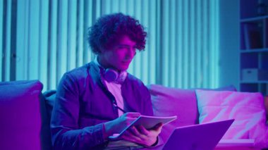 Genç Adam Neon Light 'ta, Kanepede Gece Çalışır, Not Defteri ve Laptop Tutar. Sakin Üretim ve Ruh hali Işığı ile Gen Z Çalışma Rutini. Uzaktan Öğrenme, Dijital Günlük, Estetik Odaklanma.