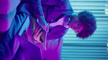 Genç Adam Neon Light 'ta, Kanepede Gece Çalışır, Not Defteri ve Laptop Tutar. Sakin Üretkenlikle Gen Z Çalışma Programı. Dikey Video, Uzaktan Öğrenme, Dijital Günlük, Estetik Odaklanma.