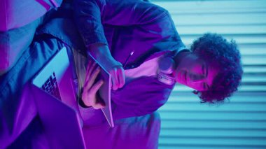 Genç Adam Neon Light 'ta, Kanepede Gece Çalışır, Not Defteri ve Laptop Tutar. Sakin Üretkenlikle Gen Z Çalışma Programı. Dikey Video, Uzaktan Öğrenme, Dijital Günlük, Estetik Odaklanma.