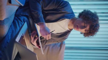 Young Man Studies at Night on Sofa in Neon Light, Holding Notebook and Laptop. Gen Z Study Routine with Calm Productivity. Vertical Video, Remote Learning, Digital Journaling, Aesthetic Focus.