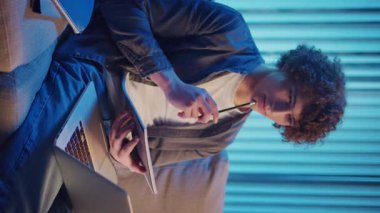 Genç Adam Neon Light 'ta, Kanepede Gece Çalışır, Not Defteri ve Laptop Tutar. Sakin Üretkenlikle Gen Z Çalışma Programı. Dikey Video, Uzaktan Öğrenme, Dijital Günlük, Estetik Odaklanma.