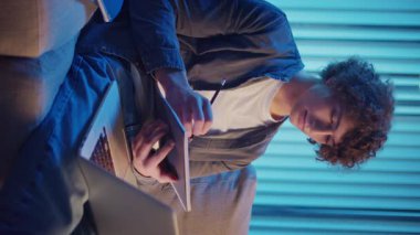 Genç Adam Neon Light 'ta, Kanepede Gece Çalışır, Not Defteri ve Laptop Tutar. Sakin Üretkenlikle Gen Z Çalışma Programı. Dikey Video, Uzaktan Öğrenme, Dijital Günlük, Estetik Odaklanma.