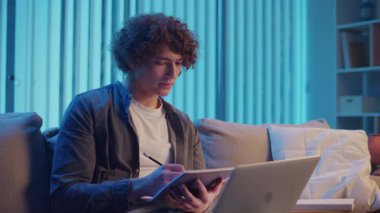 Genç Adam Neon Light 'ta, Kanepede Gece Çalışır, Not Defteri ve Laptop Tutar. Sakin Üretim ve Ruh hali Işığı ile Gen Z Çalışma Rutini. Uzaktan Öğrenme, Dijital Günlük, Estetik Odaklanma.