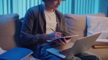 Genç Adam Neon Light 'ta, Kanepede Gece Çalışır, Not Defteri ve Laptop Tutar. Sakin Üretim ve Ruh hali Işığı ile Gen Z Çalışma Rutini. Uzaktan Öğrenme, Dijital Günlük, Estetik Odaklanma.