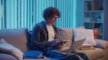 Genç Adam Neon Light 'ta, Kanepede Gece Çalışır, Not Defteri ve Laptop Tutar. Sakin Üretim ve Ruh hali Işığı ile Gen Z Çalışma Rutini. Uzaktan Öğrenme, Dijital Günlük, Estetik Odaklanma.