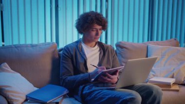 Genç Adam Neon Light 'ta, Kanepede Gece Çalışır, Not Defteri ve Laptop Tutar. Sakin Üretim ve Ruh hali Işığı ile Gen Z Çalışma Rutini. Uzaktan Öğrenme, Dijital Günlük, Estetik Odaklanma.