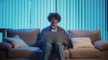 Genç Adam geceleri koltukta dizüstü bilgisayarla Kucak 'ta oturur, Online bir toplantıya katılır. Melez Yaşam Tarzında Sıradan Ama Üretken Bir Gece. Uzaktan Çalışma, Dijital İletişim, Odaklanma Kipi.