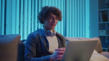 Genç Adam geceleri koltukta dizüstü bilgisayarla Kucak 'ta oturur, Online bir toplantıya katılır. Melez Yaşam Tarzında Sıradan Ama Üretken Bir Gece. Uzaktan Çalışma, Dijital İletişim, Odaklanma Kipi.