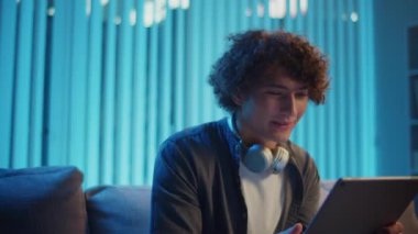 Genç Adam geceleri koltukta dizüstü bilgisayarla Kucak 'ta oturur, Online bir toplantıya katılır. Melez Yaşam Tarzında Sıradan Ama Üretken Bir Gece. Uzaktan Çalışma, Dijital İletişim, Odaklanma Kipi.