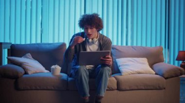 Genç Adam Tablet ve Patlamış Mısırla Gece Kanepesinde Oturuyor, Videonun tadını çıkarıyor. Rahatlama ve kişisel zaman vasıtasıyla Akşam Kişisel Bakım. Dijital Boş Zaman, Duygusal Yeniden Yükleme, Ekran Eğlencesi.