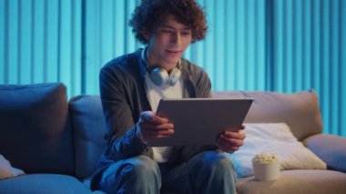 Young Man Sits on Sofa at Night with Tablet and Popcorn, Enjoying a Video. Evening Self-Care Through Relaxation and Personal Time. Digital Leisure, Emotional Recharge, Screen Entertainment.