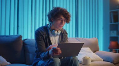 Young Man Sits on Sofa at Night with Tablet and Popcorn, Enjoying a Video. Evening Self-Care Through Relaxation and Personal Time. Digital Leisure, Emotional Recharge, Screen Entertainment.