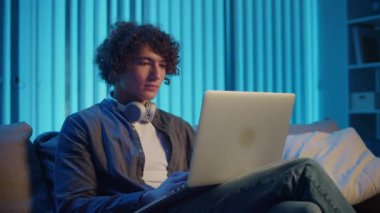 Genç adam geceleri odasında koltuğunda oturur, çalışır ya da dizüstü bilgisayarda çalışır. Tech-Driven Dünyasında Bir Öğrencinin Akşam Programı. Uzaktan Öğrenme, Dijital Üretim, Zihinsel Odaklanma.