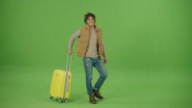Sıcak Yelek Giyen Genç Kıvırcık Meksikalı Turist Sarı Trolley Torbası, Ellerini kemerin üstüne koyup, Etrafa bakıyor ve Yeşil Ekran Arkaplanında Kazanan Hareketler Yapıyor