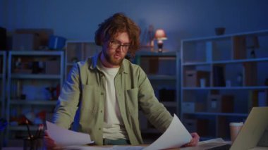 Mimari Planlama. Profesyonel bir adam dikkatle büyük planları gece geç saatlerde masasında gözden geçirir. Sahne, Mimarlık, Mühendislik, Araştırma ve Kompleks Projeler İsteklerini Sembolize eder.