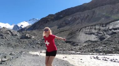 İsviçre 'nin Morteratsch buzulunun dibinde kırmızı İsviçre bayrağı tişörtü giymiş bir kadın ağır çekimde zıplıyor. İsviçre 'nin Graubunden kantonundaki Bundner Alpleri' nin Bernina Sıradağları.