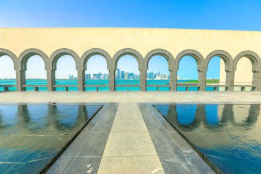 Doha, Katar-16 Şubat 2019: Islam Sanatı Müzesi veya Mia 'da kemerli yürüyüş yolu ve yansıtmalı havuzların görünümü. Doha West Bay ve Basra Körfezi arka planda. Doha Körfezi sahili. Güneşli gün.